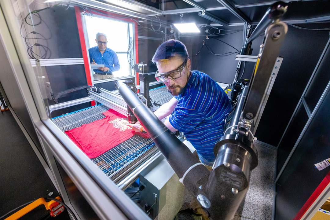 Two staff members with a robotic arm working on a red tshirt