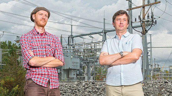 Eric Hittinger and Eric Williams post for a photo together in front of power lines, both of their arms crossed.