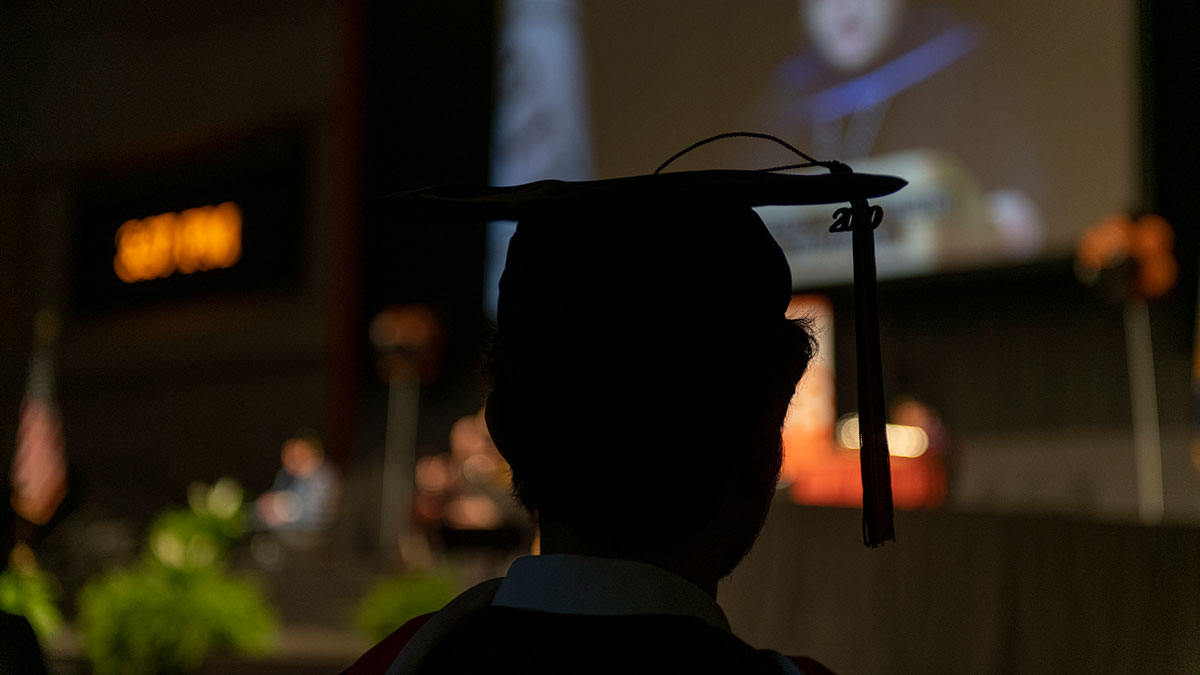 A silhouetted graduate wearing a cap and tassel watching a speaker on stage during a ceremony.