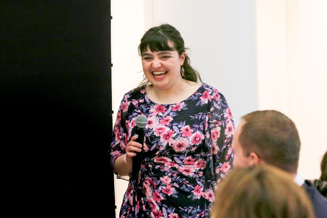 a studeny in a floral dress and long brown hair speaks in to a microphone at an event