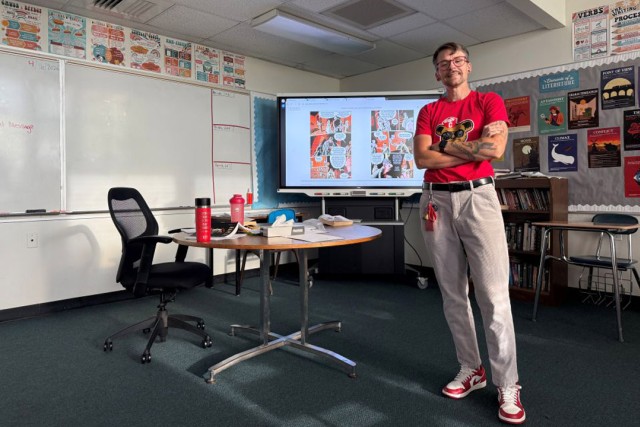a tall person wearing khaki pants and a red shirt stands in a classroom with a whiteboard lit up with a lesson behind them.