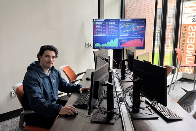 a young man with dark hair sites in front of a computer station in a lab running a bloomberg terminal