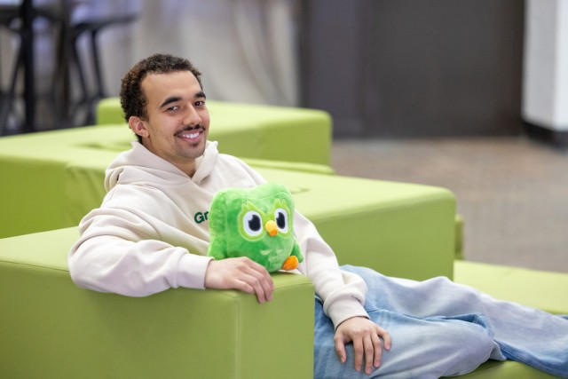 a man sits on a green chair holding a stuffed animal of the duolingo owl mascot