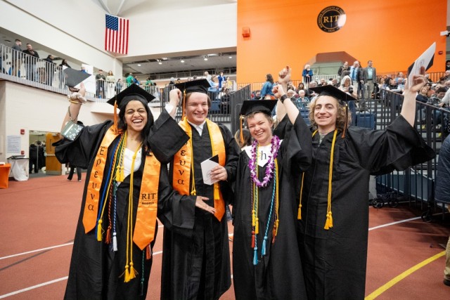 Four college graduates in an arena smile and cheer while wearing black caps and gowns.