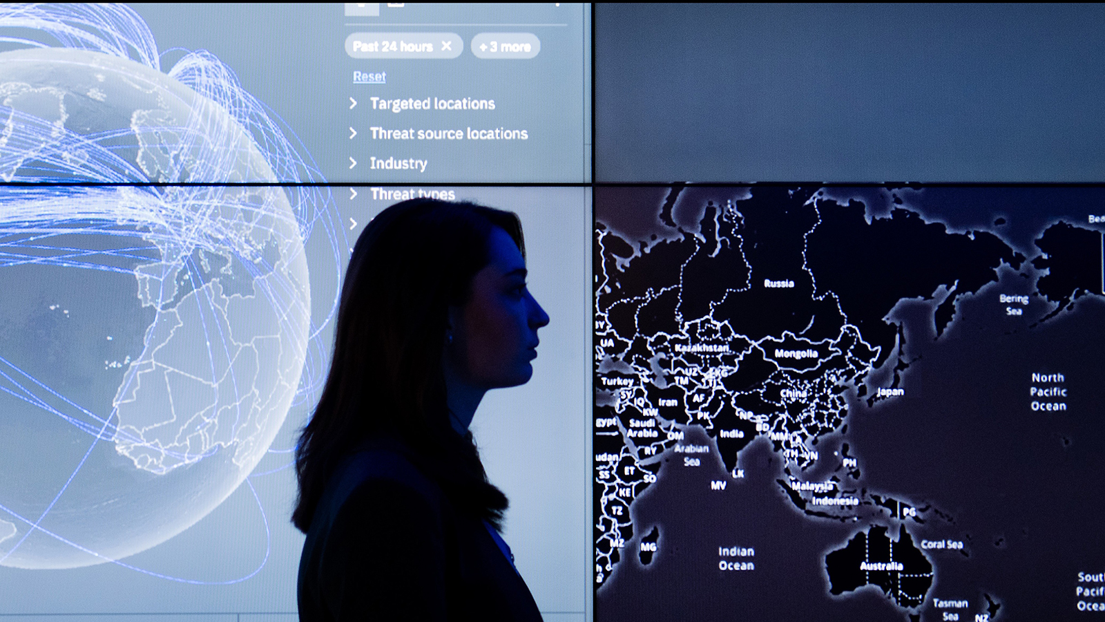 Silhouette of a person observing two large digital screens displaying global data, with one screen showing a world map highlighting various locations.