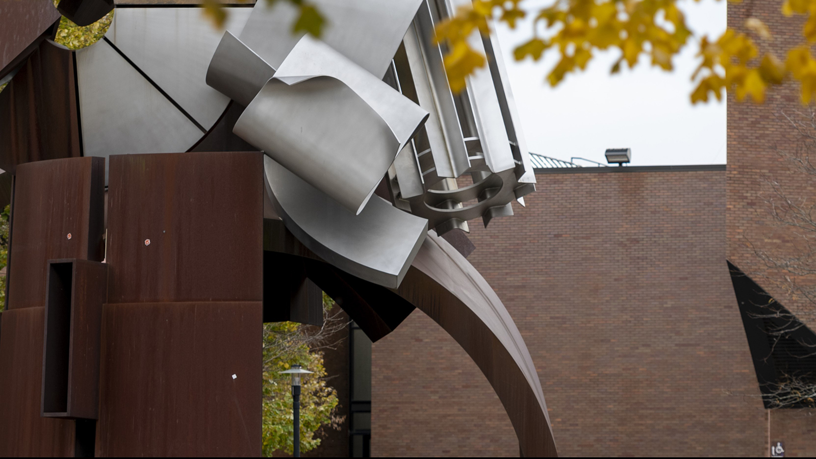 An abstract metal sculpture with geometric shapes set against autumn trees and a brick building.