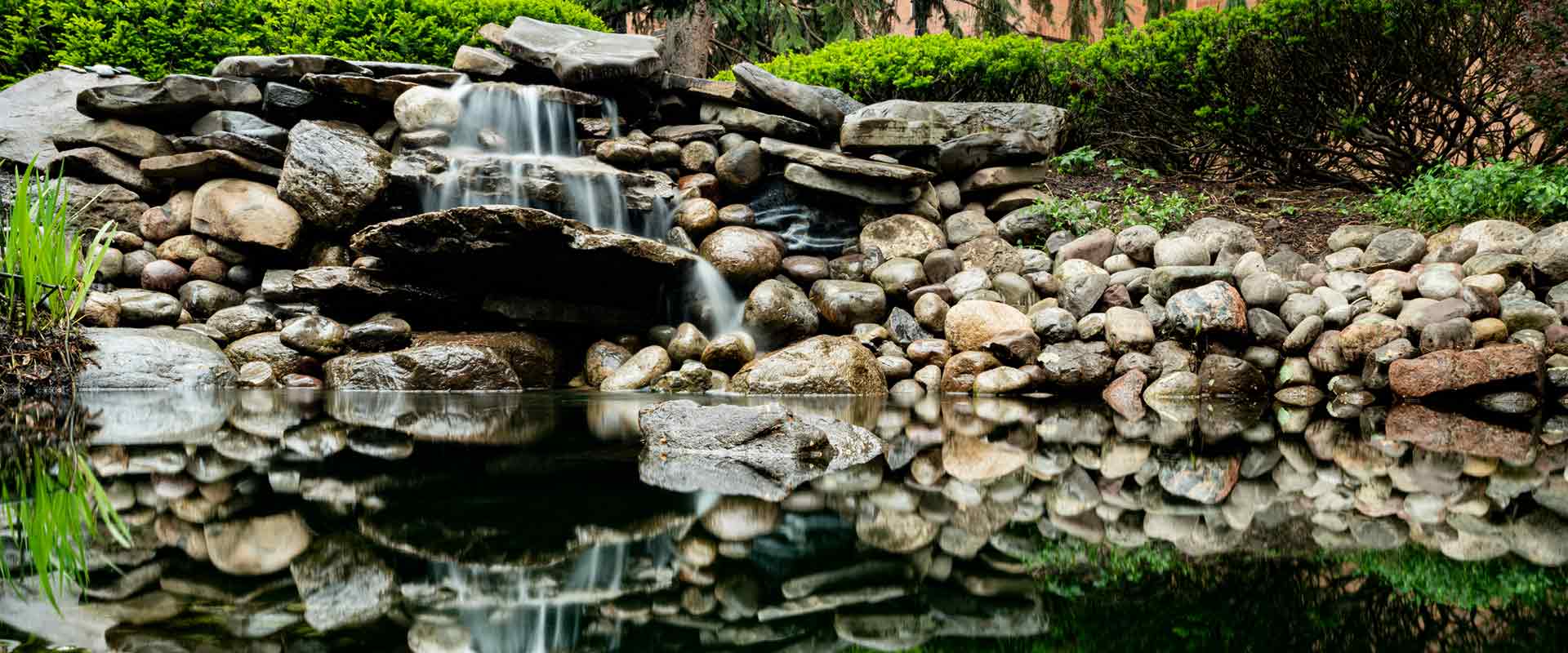 a small waterfalls spills around rocks into a calm pond, which reflects the surrounding rocks and greenery.