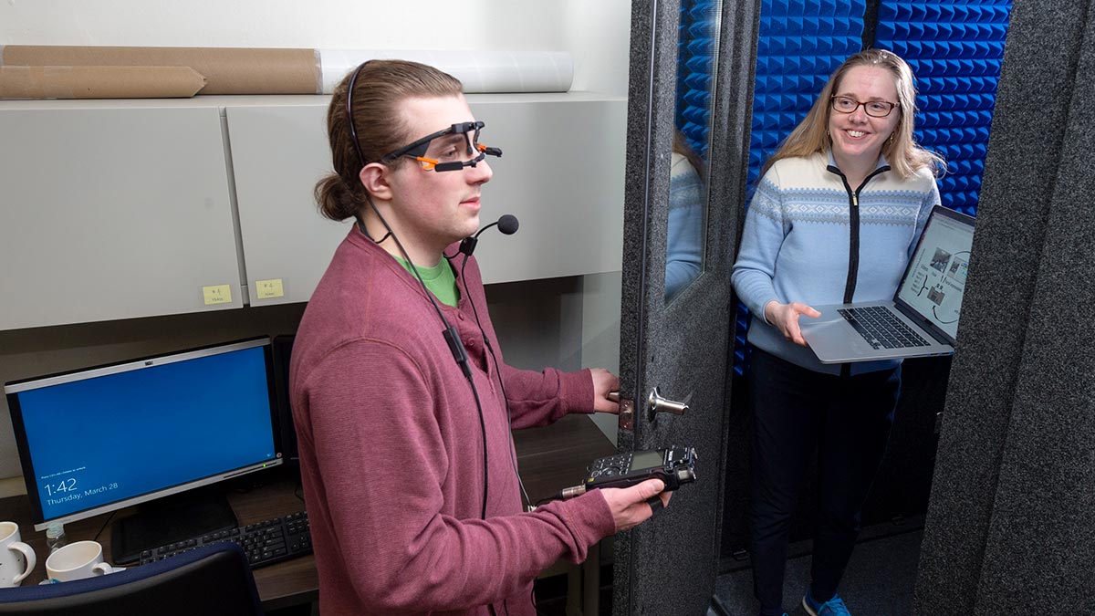 one person wearing a special headset with glasses and a microphone while another person opens the door to a sound booth.