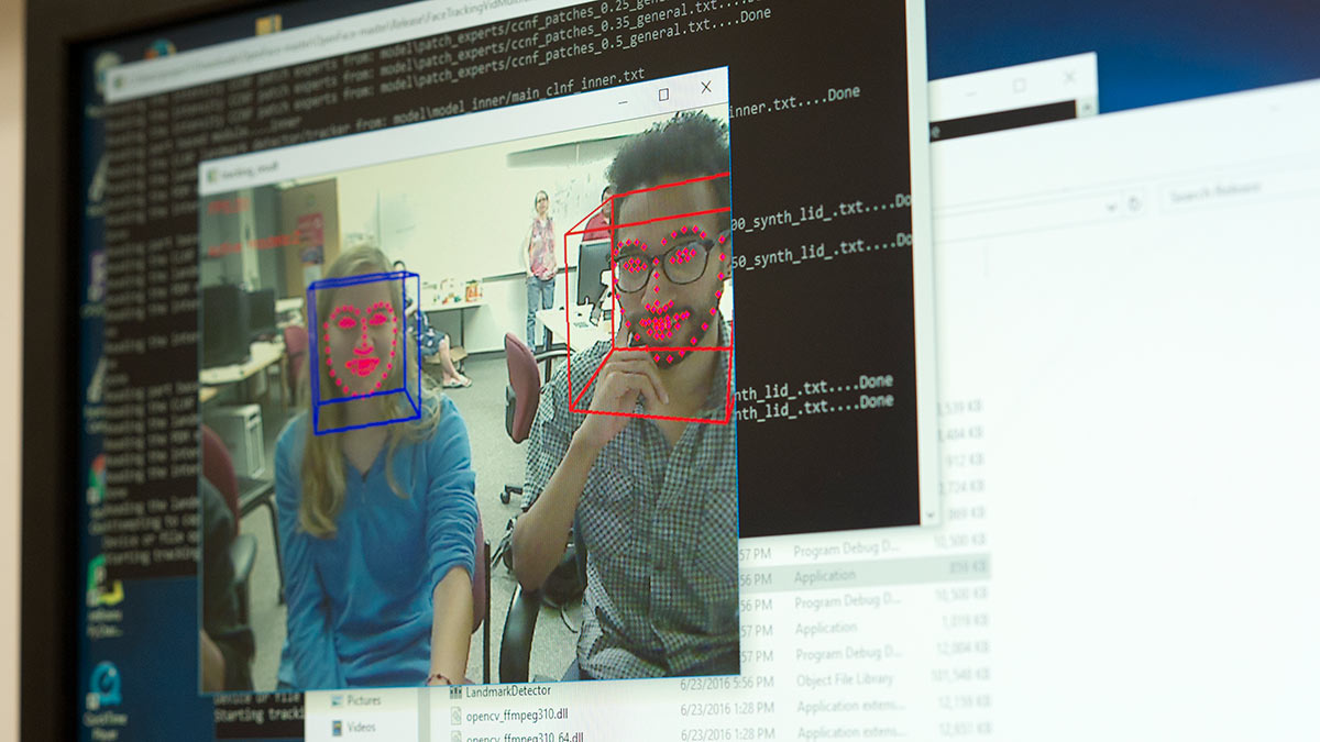 computer screen showing a video of two people with 3 D boxes around their heads and red dots outlining their face, mouths, and eyes.