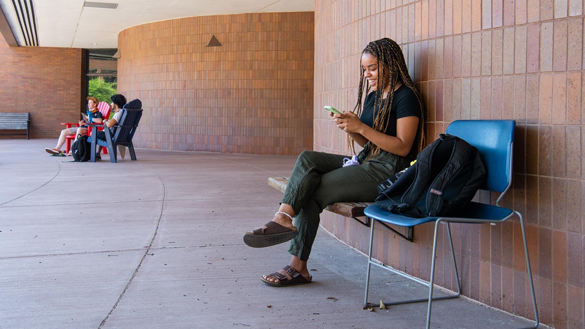 a smiling woman sits outside on a bench and looks at her phone.