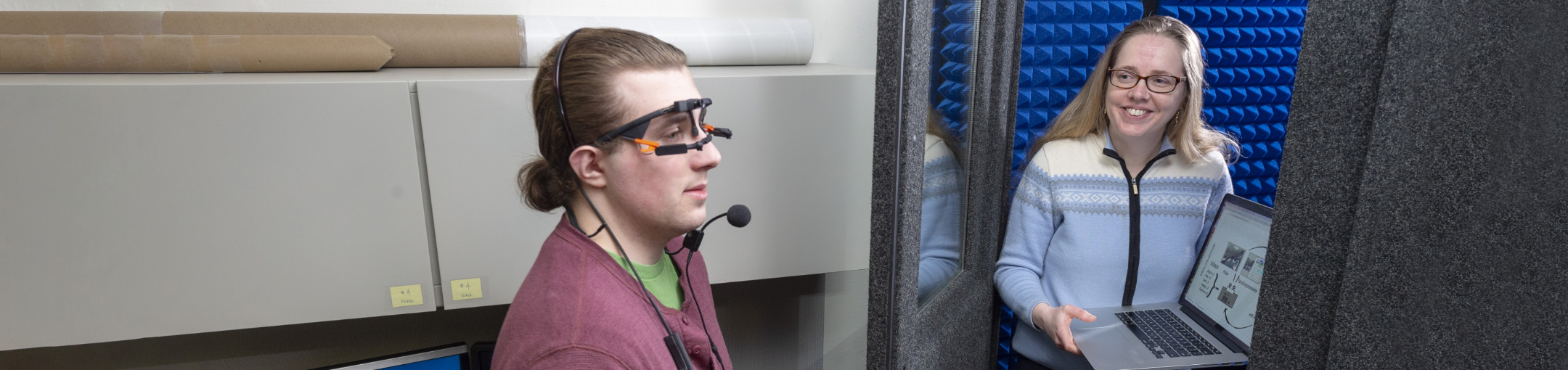 one person wearing a special headset with glasses and a microphone while another person opens the door to a sound booth.