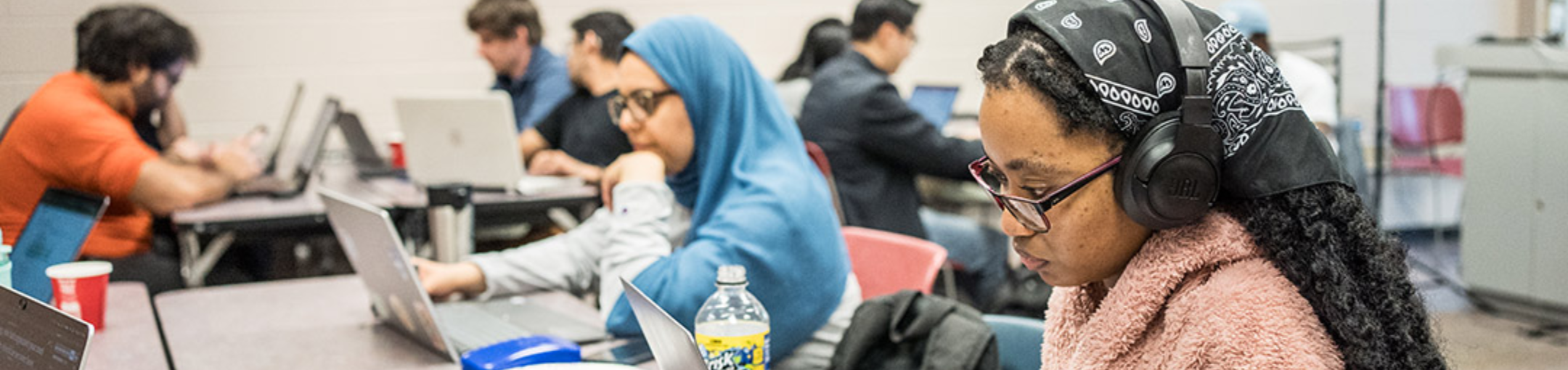 people sitting at tables on laptops. the person in the foreground is wearing head phones staring intently