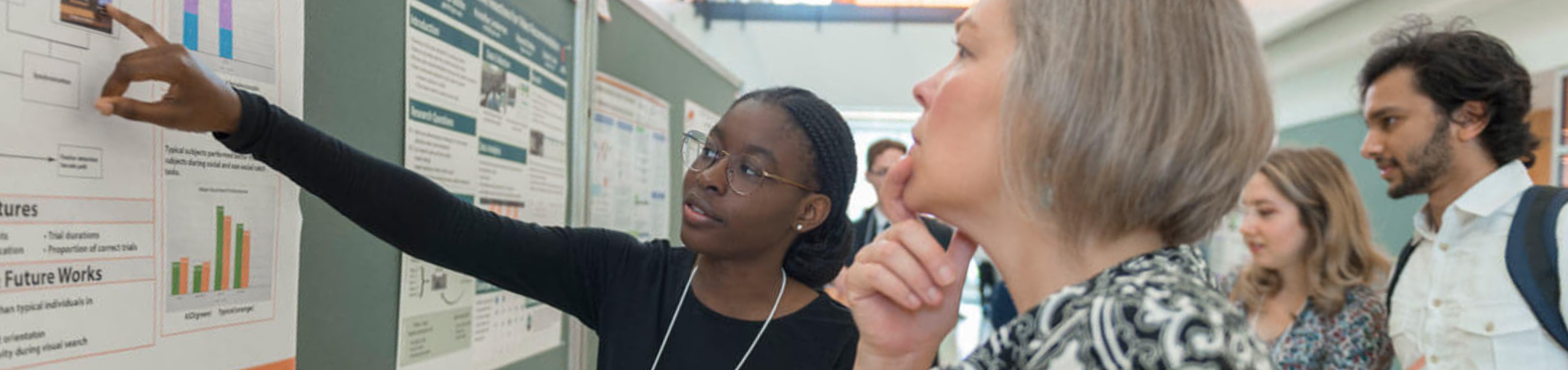 A person pointing to a chart posted on a wall while another person touches their chin thoughtfully while looking at the poster