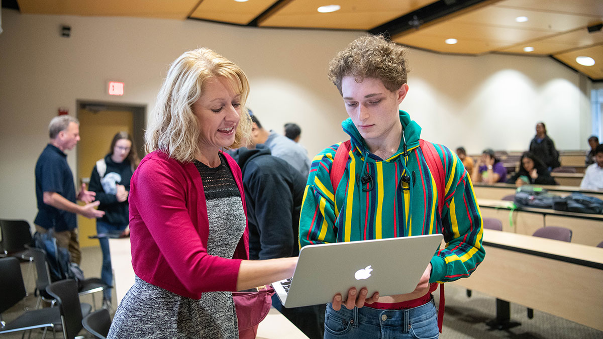 A woman assists a student with a laptop in a classroom setting.
