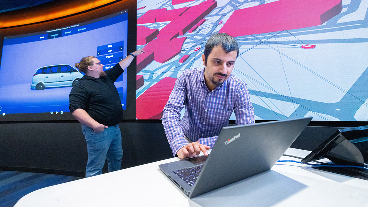 A faculty member at a laptop in the foreground, with a student pointing at 2 large screens in the background.