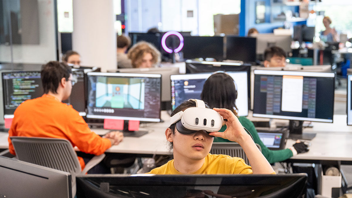 Students working on computers in a lab, with one student wearing a virtual reality headset.