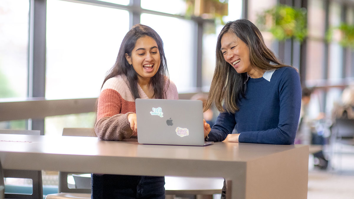 Two students smiling and discussing over a laptop in a bright, open study space with large windows.