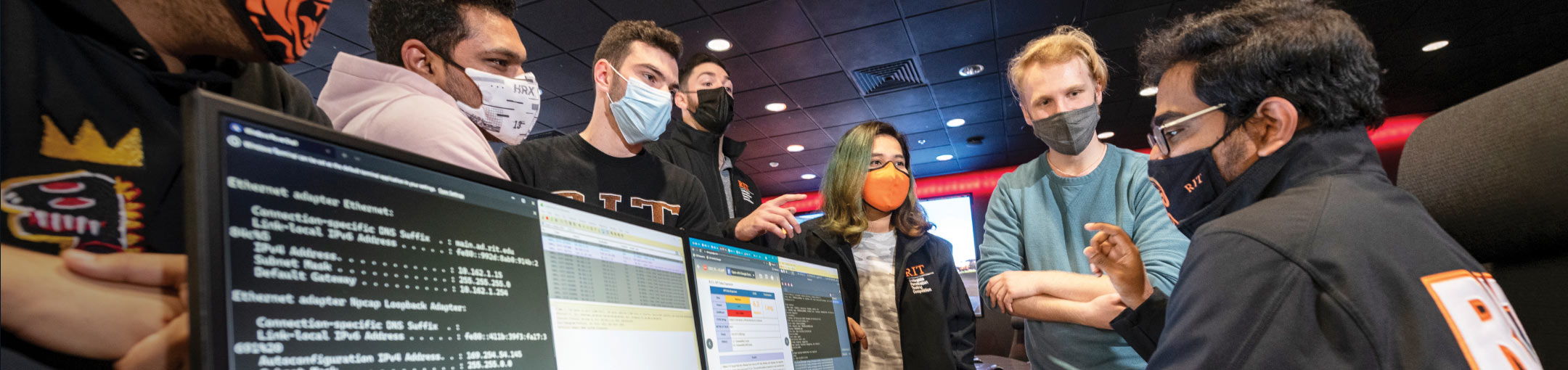 A student sits in front of multiple monitors as other students look on.