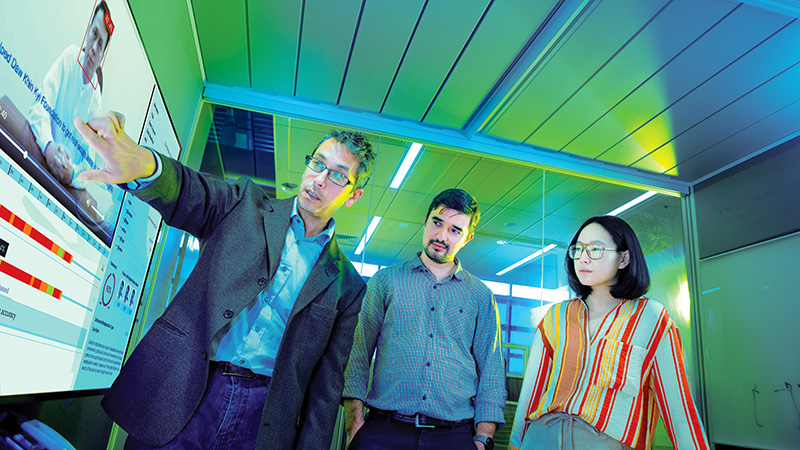 A faculty member pointing to a large screen showing analysis of a face, while 2 people are standing next to him.