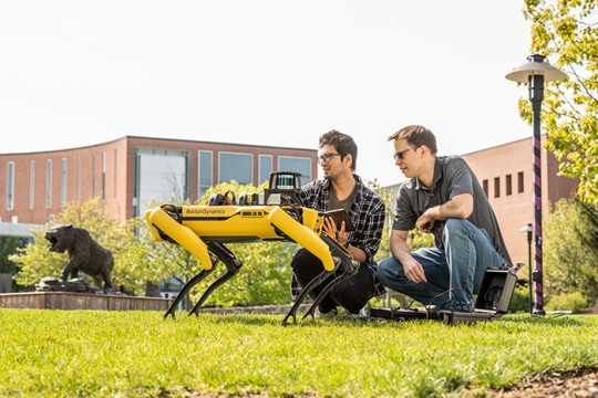 Two people squatted down, looking at Boston Dynamics robot, Spot who is yellow and shaped like a dog.