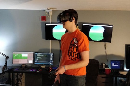 Student wearing a virtual reality headset standing in front of computer monitors.