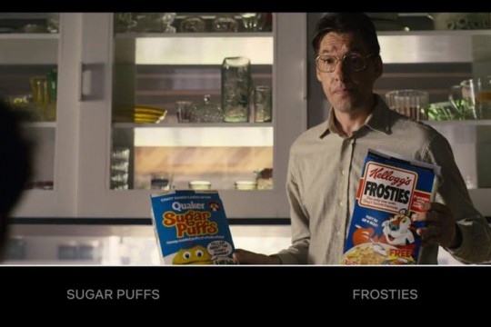 Screenshot of TV show. Man stands holding a box of Kellogg's Frosties in one hand and QUaker Sugar Puffs in the other hand
