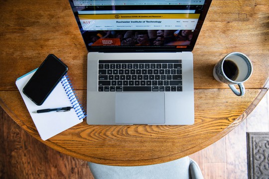 Overhead view of open laptop on kitchen table next to mug of coffee, notepad and cell phone.