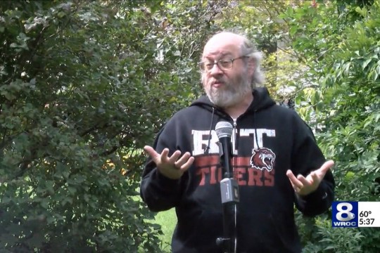 professor wearing RIT Tigers sweatshirt talking into microphone outdoors.
