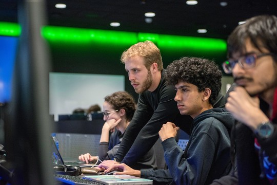 four college students lined up a table looking at computer monitors.