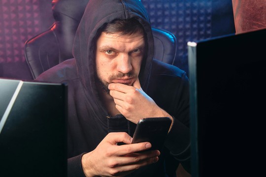 a man in a black hoodie in front of a monitor looks at a phone with a malicious look on his face