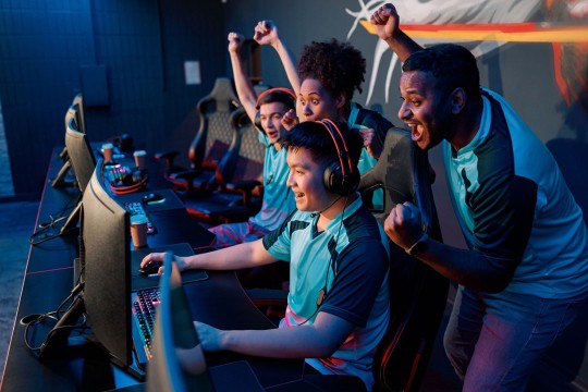 Esports team in matching jerseys celebrating a victory at a gaming tournament, with a player at a computer wearing a headset and teammates cheering behind.