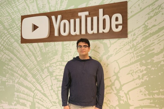 a man with dark hair and glasses stands under a YouTube sign on a green and white patterned wall.