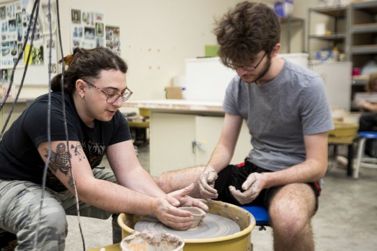 two students work together manipulating clay on a pottery wheel in a clay studio