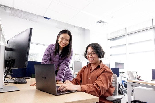 Two students sit in a modern and brightly lit computer lab, working on a laptop.