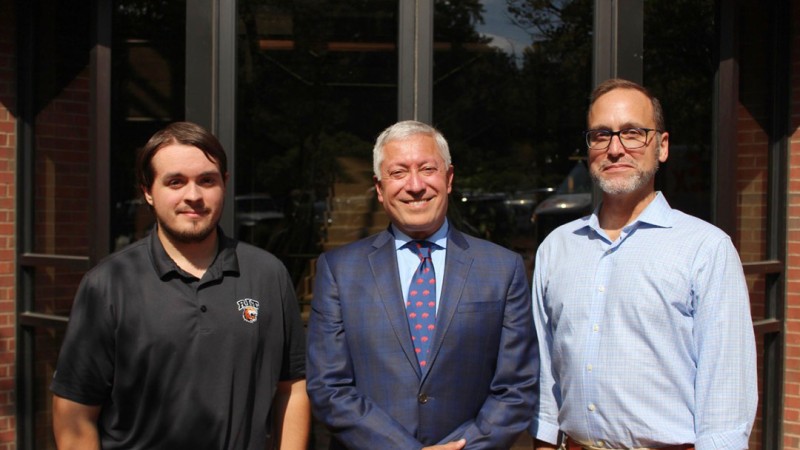 Three individuals stand in front of a glass entrance with brick framing. The person on the left is wearing a black polo shirt with a logo, the person in the center is dressed in a blue plaid suit with a patterned tie, and the person on the right is wearing a light blue button-down shirt with khaki pants and a brown belt.