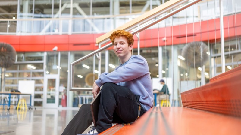 Benjamin Brundage in blue sweatshirt and black pants sits on orange stairwell in RIT SHED