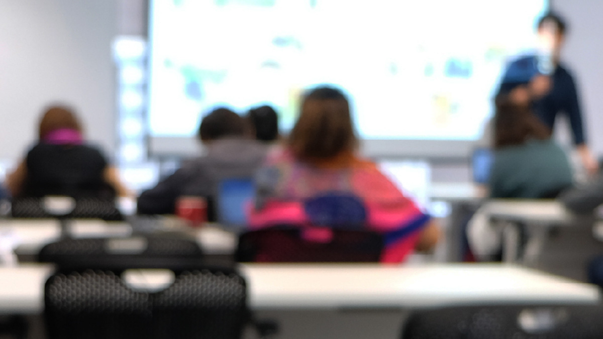 Group of people sitting at desks in a classroom with an instructor teaching at the front of the room.