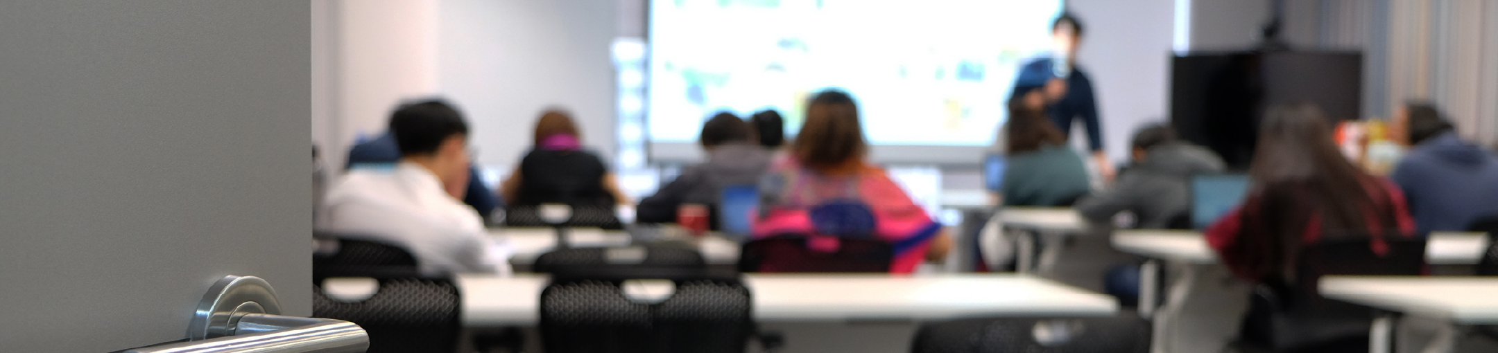 Group of people sitting at desks in a classroom with an instructor teaching at the front of the room