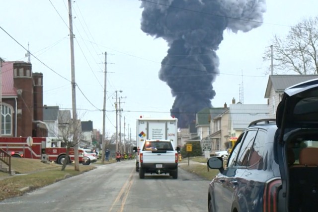 view down a neighborhood street with a plume of dark smoke in the background.