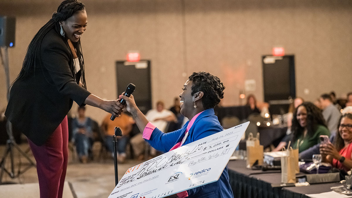a woman on a stage hands a microphone to another woman who is holding a giant novelty check.
