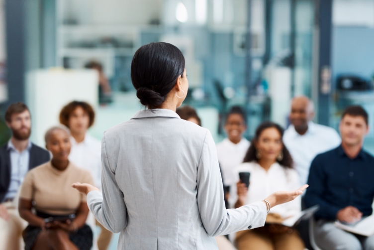 A women with her back to the camper talking to a group of people seated in business casual attire