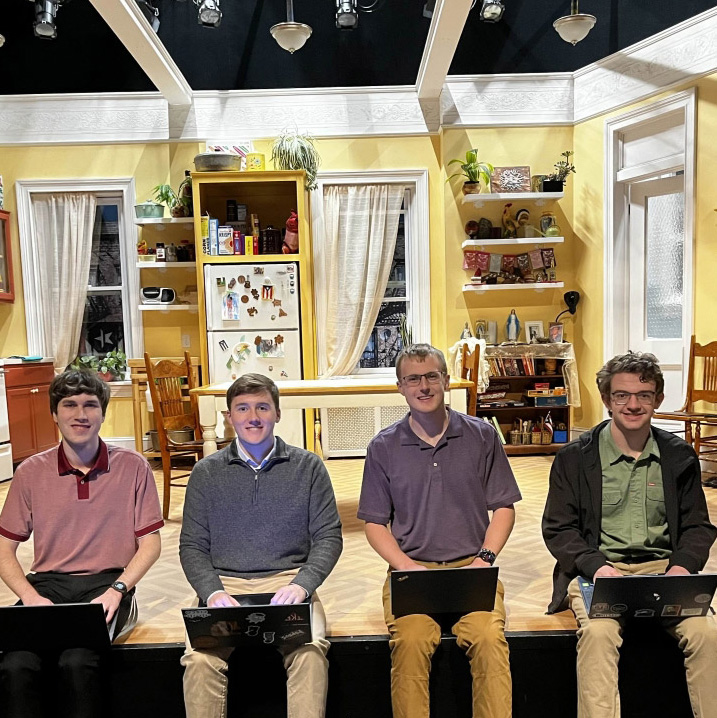 Group of 4 students with laptops on their laps sit on edge of theater stage