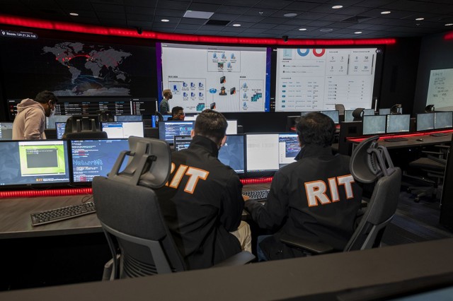 students sitting at long tables with computers and two large projector screens in front of the room.
