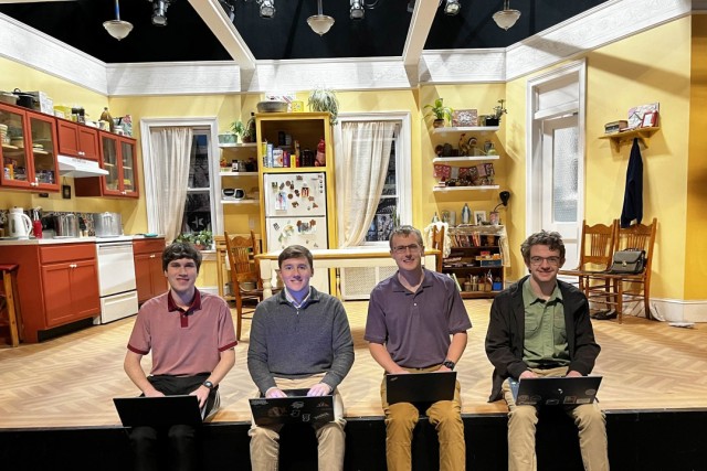 four students sit on a stage with the set for a play behind them