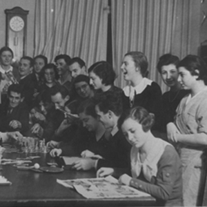 Party of people looking over the shoulder of someone sitting at a table writing