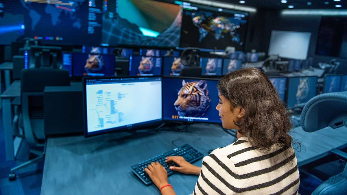 A woman working at dual monitors in a cybersecurity command center, analyzing a digital diagram with a background of multiple workstations.