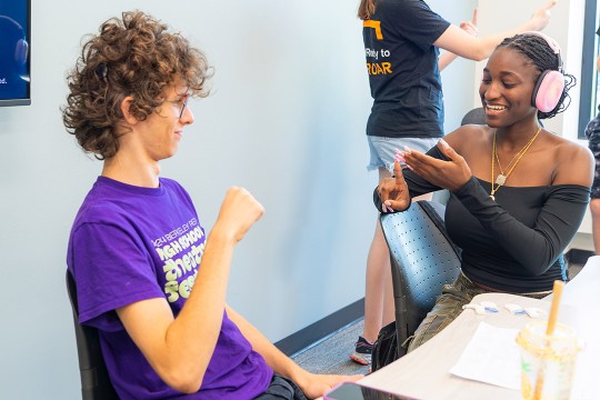 a white college age male sits next to a black college age female in headphones. They are both speaking sign language.
