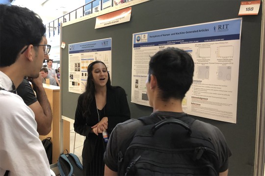 a college age woman speaks to two people as they all stand in front of a poster.