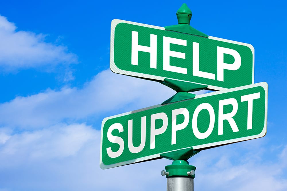 Two green street signs reading “HELP” and “SUPPORT” intersect against a bright blue sky with clouds.