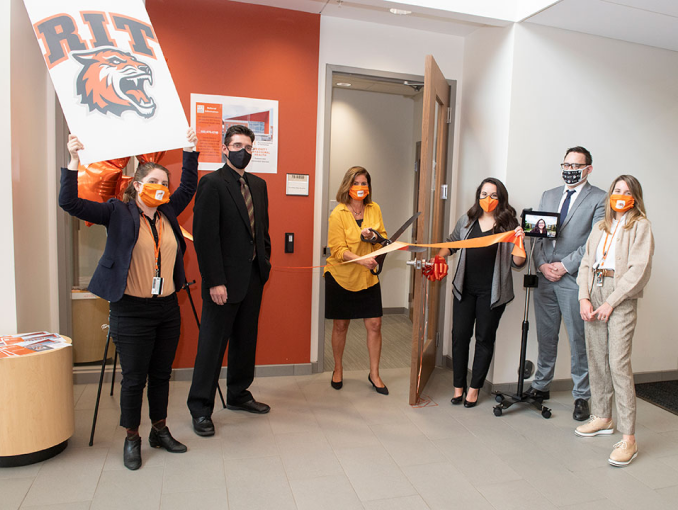 six people celebrate opening of a clinic by cutting a ribbon with oversized scissors.
